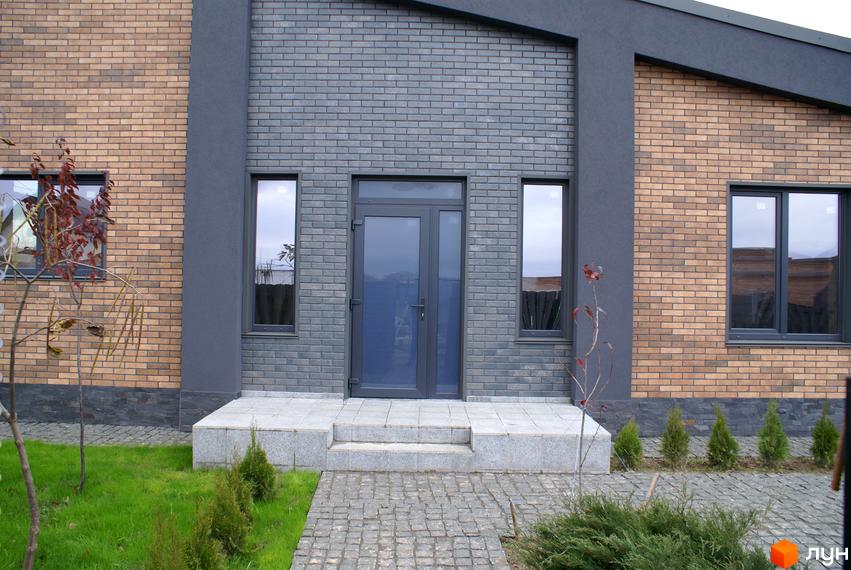 Котеджне містечко КМ Country Villa. Фасад будинку з цегли двох кольорів, сірого та бежевого, з темними вікнами та дверима. Перед будинком доглянута прибудинкова територія з газоном та доріжками.