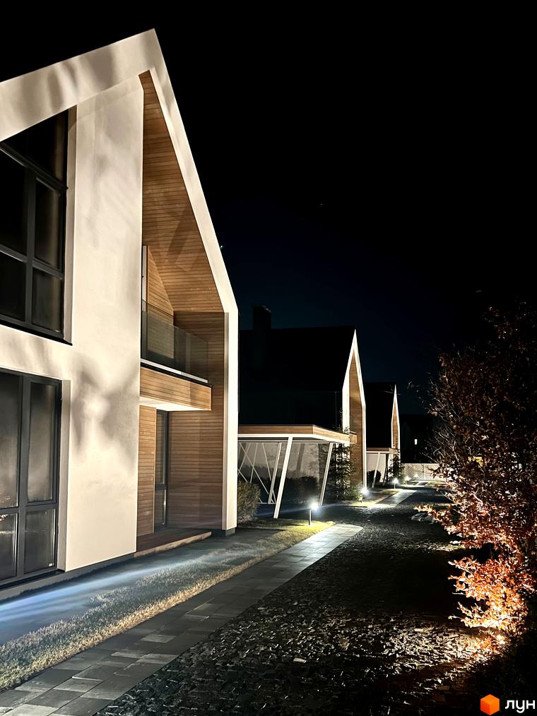 Котеджне містечко КМ Hill Park з дерев'яними елементами фасаду та підсвіченою територією вночі. Сучасна архітектура новобудови та доглянутий ландшафт.