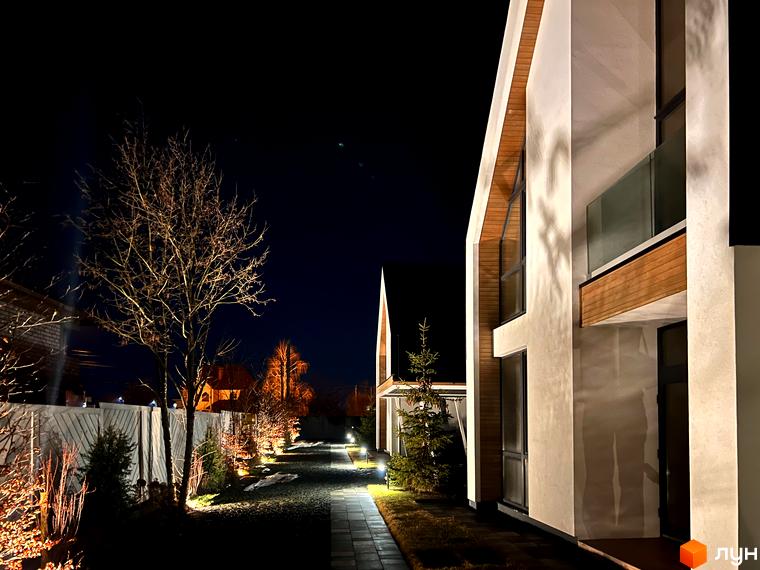 Котеджне містечко КМ Hill Park: фасади будинків з дерев'яними елементами та скляними балконами, освітлена алея з рослинами вночі.