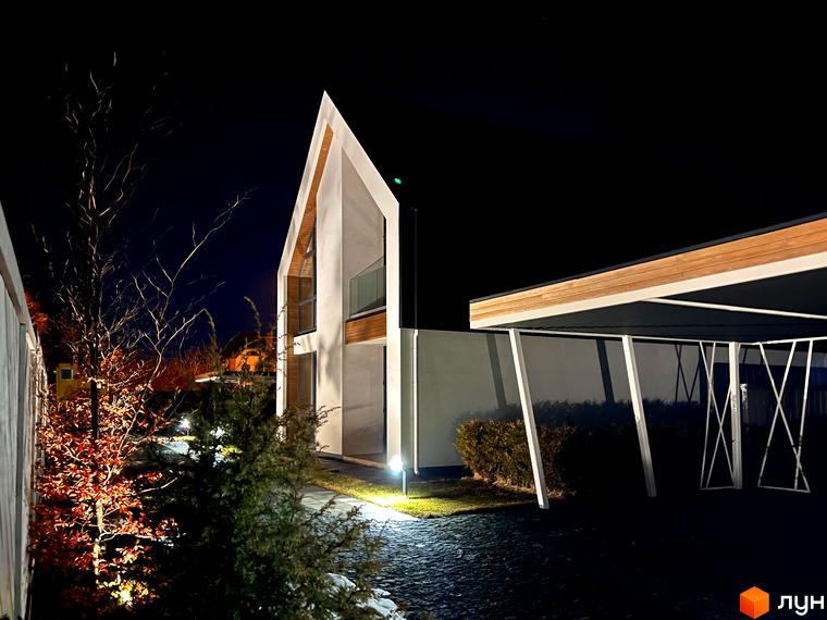 Котеджне містечко КМ Hill Park. Сучасний будинок з панорамними вікнами та навісом для авто, освітлений вночі. Прибудинкова територія з деревами та кущами.