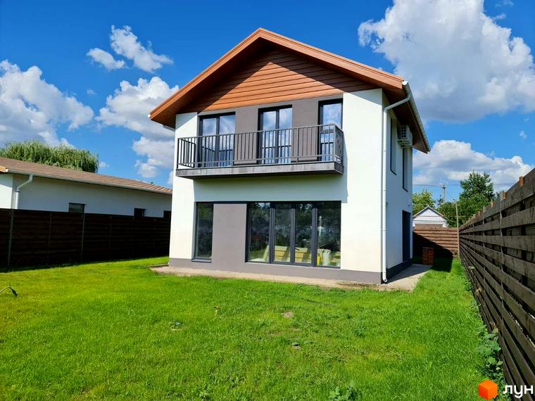 Котеджне містечко Заміський: двоповерховий будинок з балконом та великими вікнами на першому поверсі, зелена галявина та дерев'яний паркан. Навколо будинку доглянута прибудинкова територія.