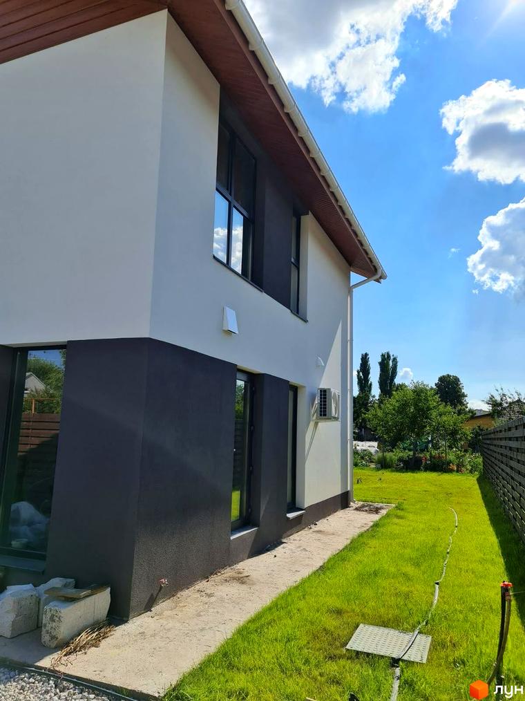 Котеджне містечко КМ Заміський: двоповерховий будинок з білою та темною штукатуркою, великими вікнами та зеленим газоном навколо. На території розташовані дерева та паркан.