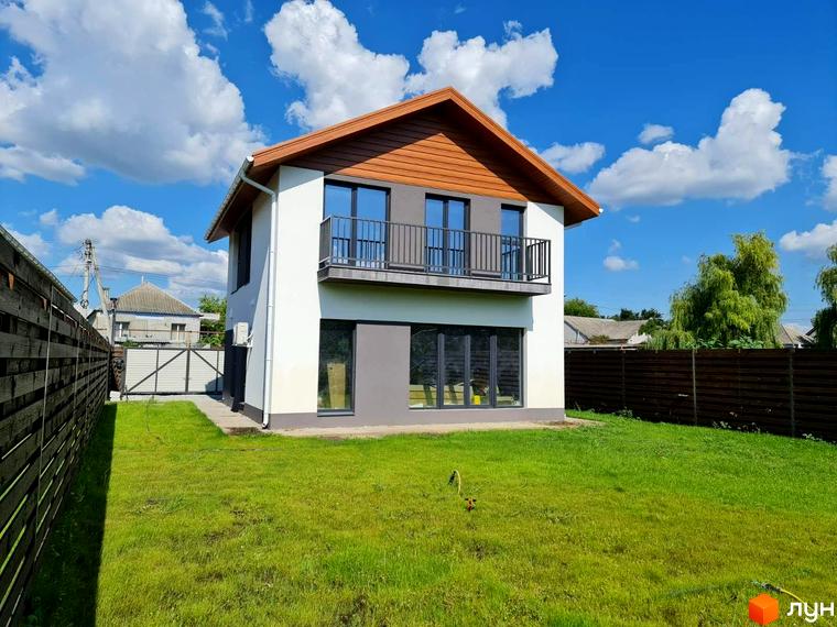 Котеджне містечко Заміський: двоповерховий будинок з балконом та великими вікнами на тлі блакитного неба з хмарами. Зелена галявина та паркан навколо території.