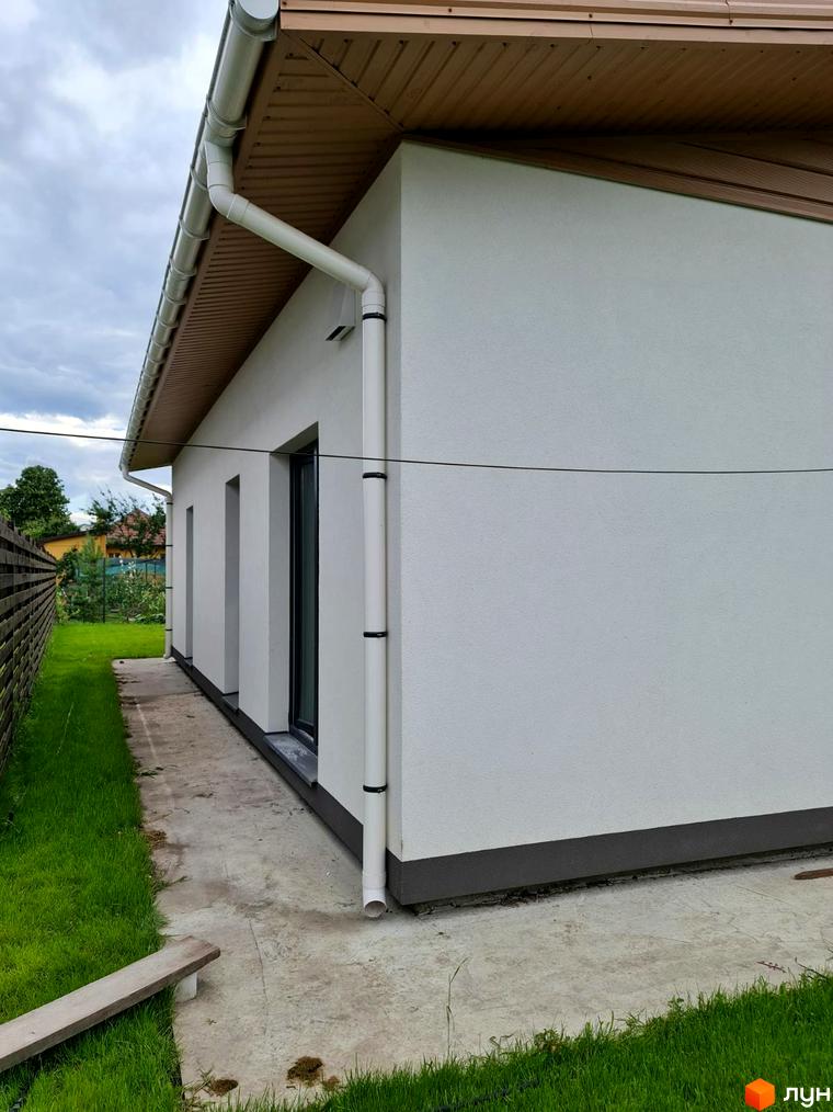 Зовнішній вигляд будинку КМ Заміський з білою стіною та білою водостічною системою. Прибудинкова територія з зеленою травою та бетонною доріжкою.