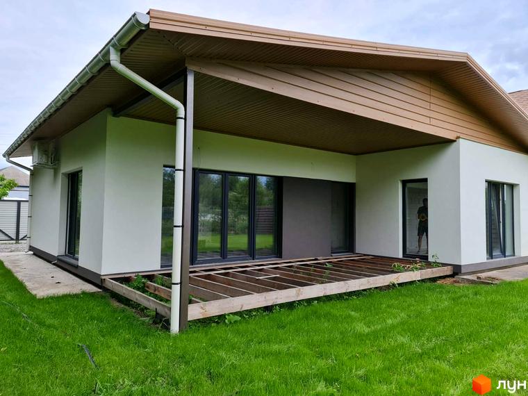 Котеджне містечко КМ Заміський: одноповерховий будинок з коричневим дахом, великими вікнами та недобудованою терасою. Навколо будинку зелена галявина та паркан.