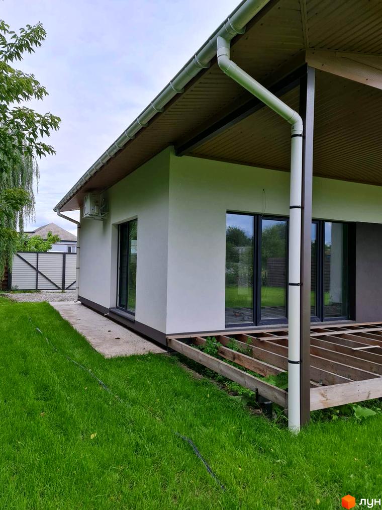 Зовнішній вигляд будинку з білою стіною, великими вікнами та дерев'яною терасою в КМ Заміський. Навколо будинку зелений газон та паркан.