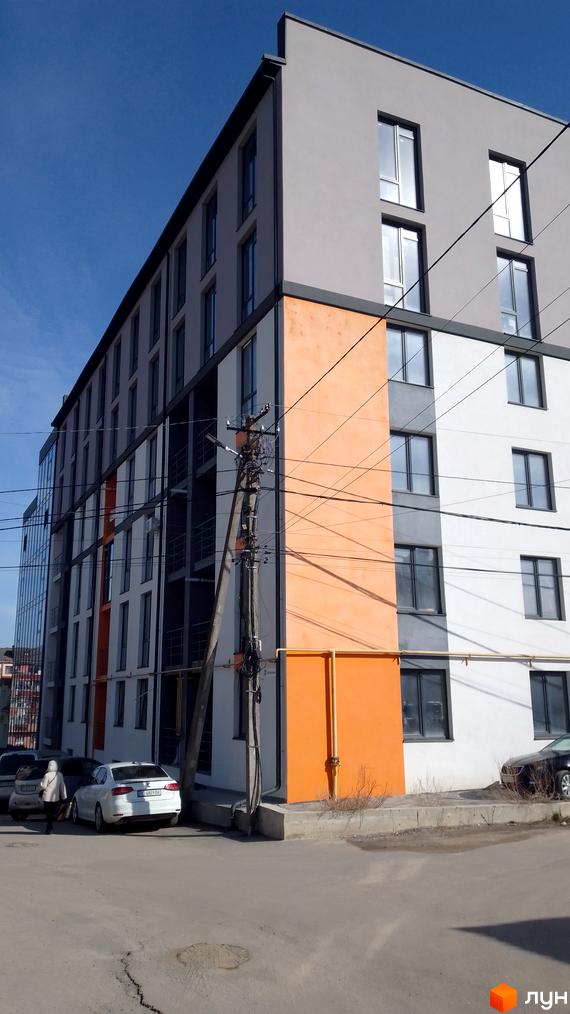 Фасад новобудови на вулиці Петриківській, 9, з сірими та білими секціями та помаранчевими акцентами. Поруч розташовані припарковані автомобілі та елементи вуличної інфраструктури.