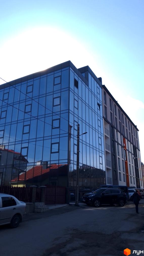 Фасад новобудови на вулиці Петриківській, 9, з великою кількістю скла та балконами. Поруч розташовані припарковані автомобілі та огорожа.