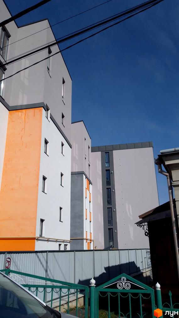 Зовнішній вигляд житлового комплексу на вул. Петриківська, 9, з білими та помаранчевими фасадами, сірими елементами та темними вікнами. На передньому плані - металева огорожа та зелені ворота.