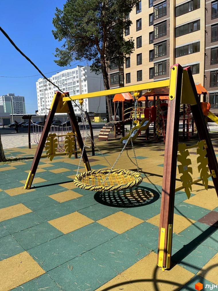 Дитячий майданчик з гойдалкою-гніздом на тлі багатоповерхових будинків ЖК Масани. Зелено-жовте покриття майданчика та дерева створюють приємне оточення.