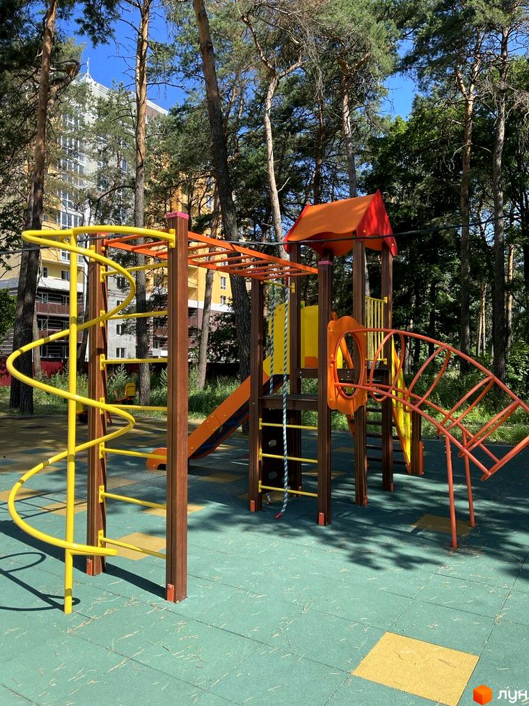 Дитячий майданчик з яскравими елементами та гірками біля багатоповерхового будинку ЖК Масани. Територія навколо майданчика вкрита зеленим покриттям.