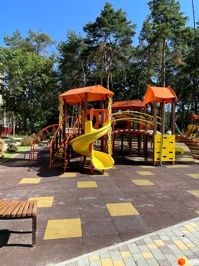 Дитячий майданчик з яскравими гірками та безпечним покриттям у житловому комплексі Масани. Навколо зелені дерева та блакитне небо.