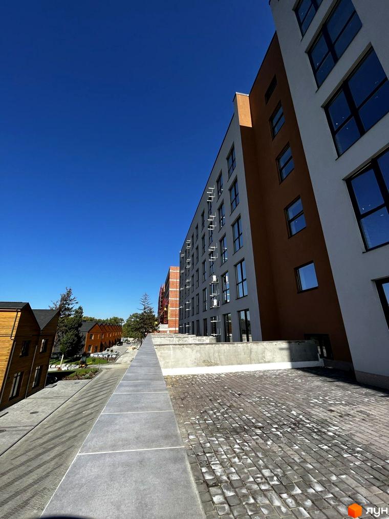 Житловий комплекс Comfort City Lagoon з фасадами біло-коричневого кольору та великими вікнами. Навколо розташовані дерев'яні будиночки та бруківка.