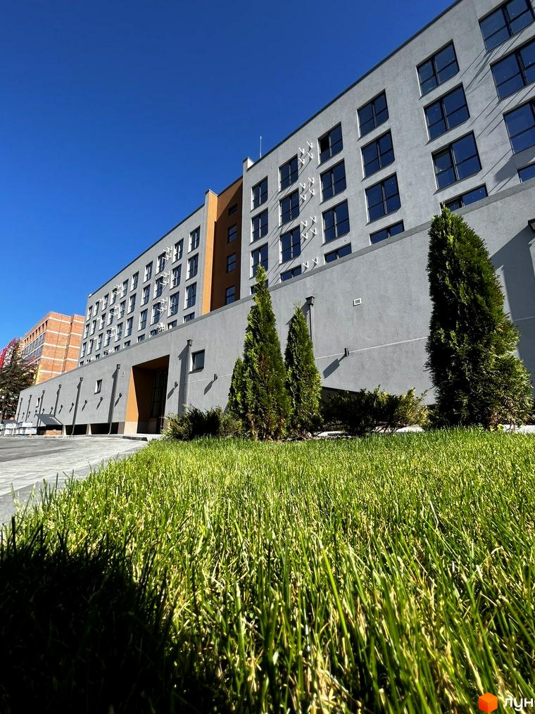 Будинок житлового комплексу Comfort City Lagoon з сірим фасадом та великими вікнами на тлі блакитного неба. Поруч розташований зелений газон та дерева.