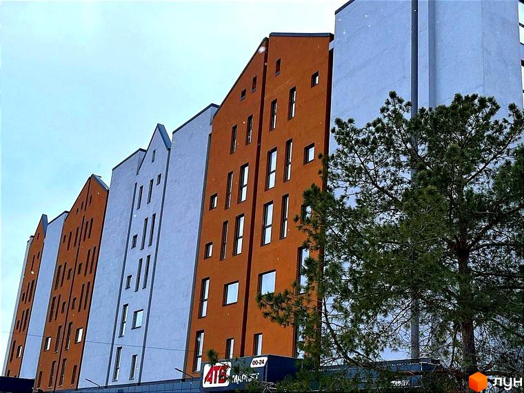 Фасад житлового комплексу Comfort City Lagoon з помаранчевими та сірими секціями, вікнами та зеленою хвоєю на передньому плані.