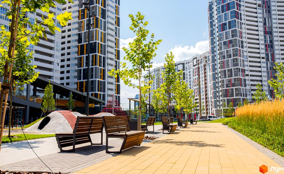Високі будинки житлового комплексу Русанівська Гавань з фасадами у світлих тонах. На території є лави, дерева та дитячий майданчик.