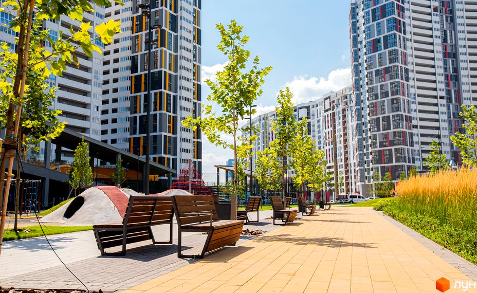 Багатоповерхові будинки Русанівська Гавань з білими, сірими та жовтими фасадами. Прибудинкова територія має доріжку, дерева та лавки.