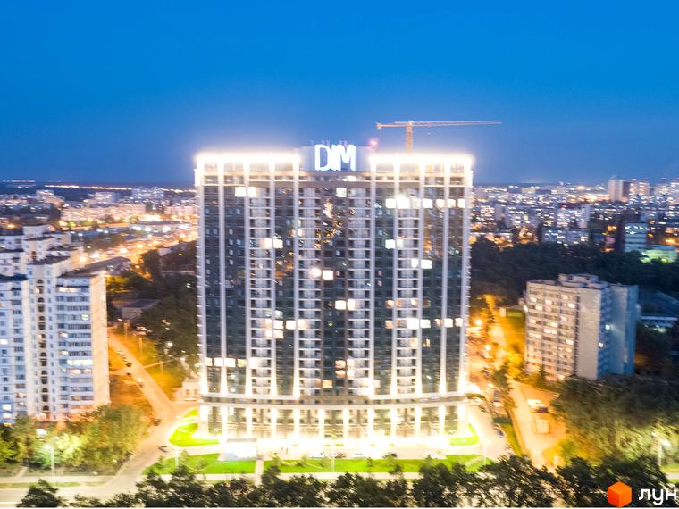 Нічний вид на новобудову ЖК Автограф з підсвіченим фасадом та міським пейзажем на задньому плані. Навколо комплексу розташовані дерева та освітлені вулиці.