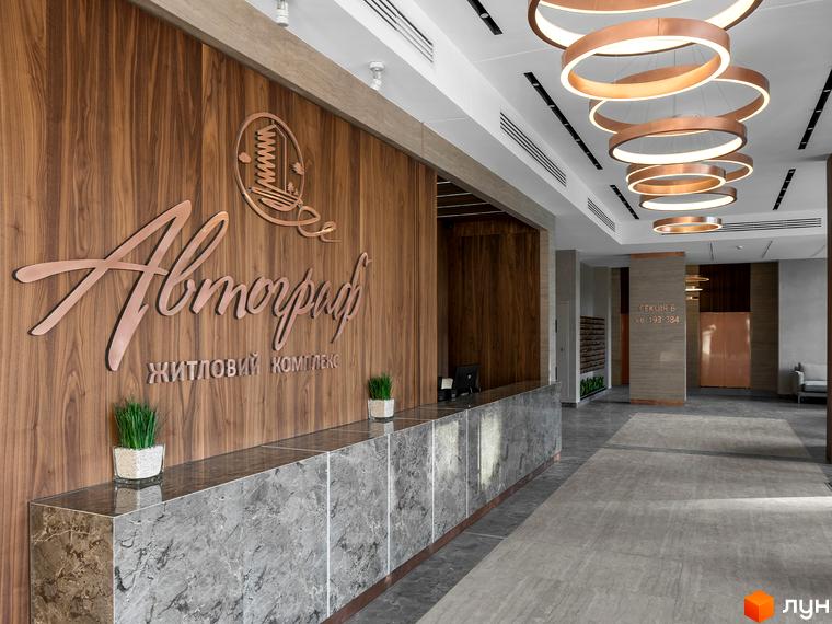 Лобі житлового комплексу Автограф з мармуровою стійкою реєстрації та дерев'яною обшивкою стін. Над стійкою розташований логотип комплексу. У стелі підвішені круглі світильники.