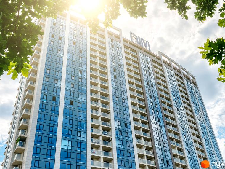 Високоякісна новобудова ЖК Автограф з панорамними вікнами та балконами, оточена зеленню та яскравим сонячним світлом. Зовнішній вигляд будівлі вражає сучасною архітектурою.