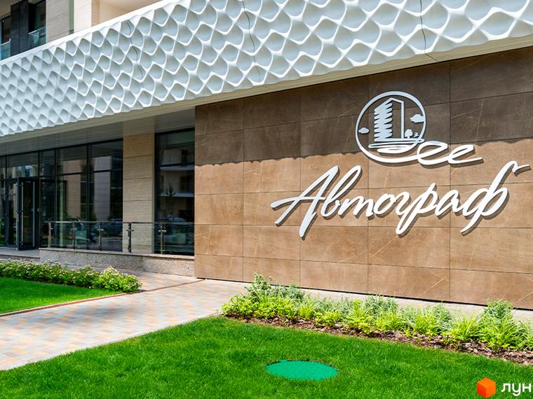 Зовнішній вигляд новобудови ЖК Автограф з коричневим фасадом та білим декоративним елементом. Прибудинкова територія з доглянутим газоном та доріжками.