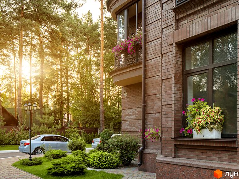 Житловий комплекс Соборний: цегляний будинок з балконом, прикрашеним квітами, та вікнами з рослинами на підвіконні. На прибудинковій території ростуть дерева та кущі, біля будинку припарковані автомобілі.