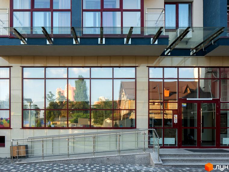Фасад новобудови ЖК Фамільний з великими вікнами та скляним навісом. Поруч розташована прибудинкова територія з пандусом та бруківкою.