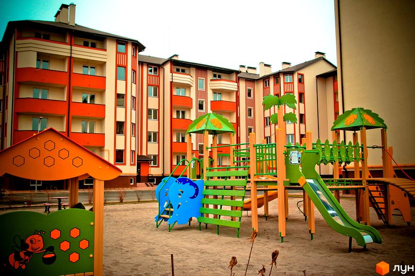 Житловий комплекс Щасливий: сучасна архітектура будівель з помаранчевими та бежевими фасадами. На прибудинковій території дитячий майданчик з гірками та гойдалками.