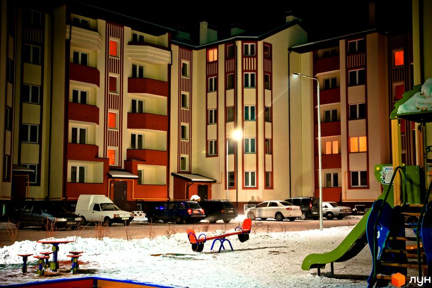 Житловий комплекс Щасливий. Багатоквартирний будинок з червоними та білими фасадами, балконами, вікнами. Перед будинком дитячий майданчик та автомобілі на парковці.