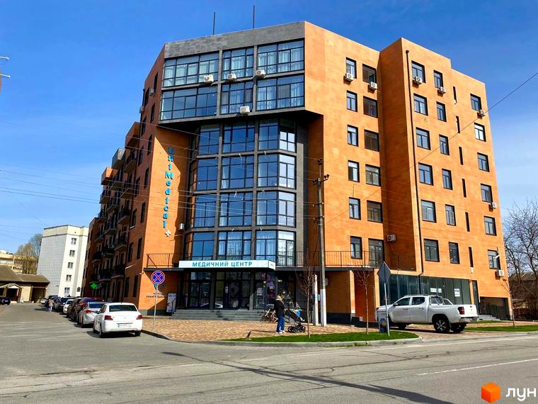 Будинок ЖК Plaza Kvartal з цегляним фасадом та великими вікнами. Поруч парковка з автомобілями та зелена зона.