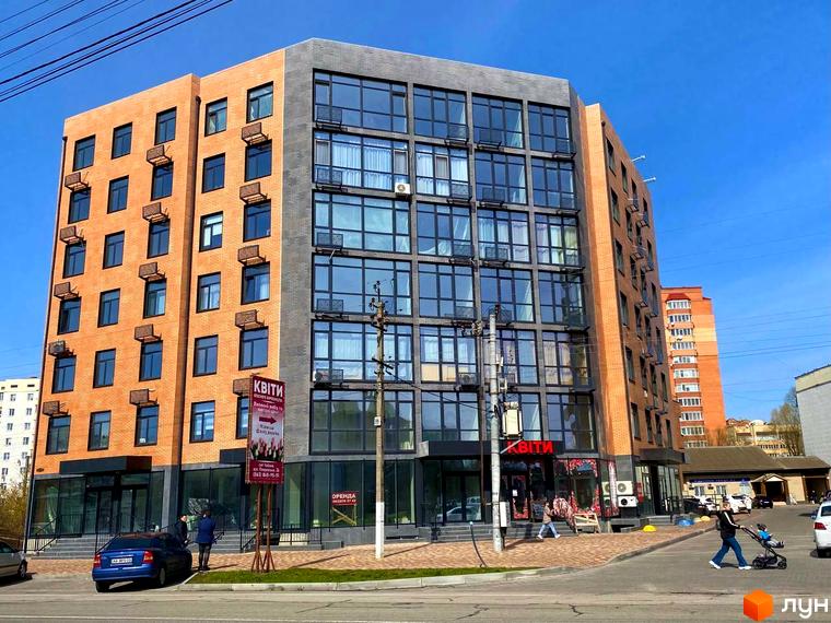 Зовнішній вигляд новобудови Plaza Kvartal з цегляним та сірим фасадом, великими вікнами та кондиціонерами. На прибудинковій території видно дорогу, тротуар та людей.