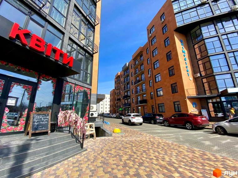 Зовнішній вигляд новобудови ЖК Plaza Kvartal з цегляним фасадом та великими вікнами. Поруч розташовані автомобілі та вимощений бруківкою прохід.