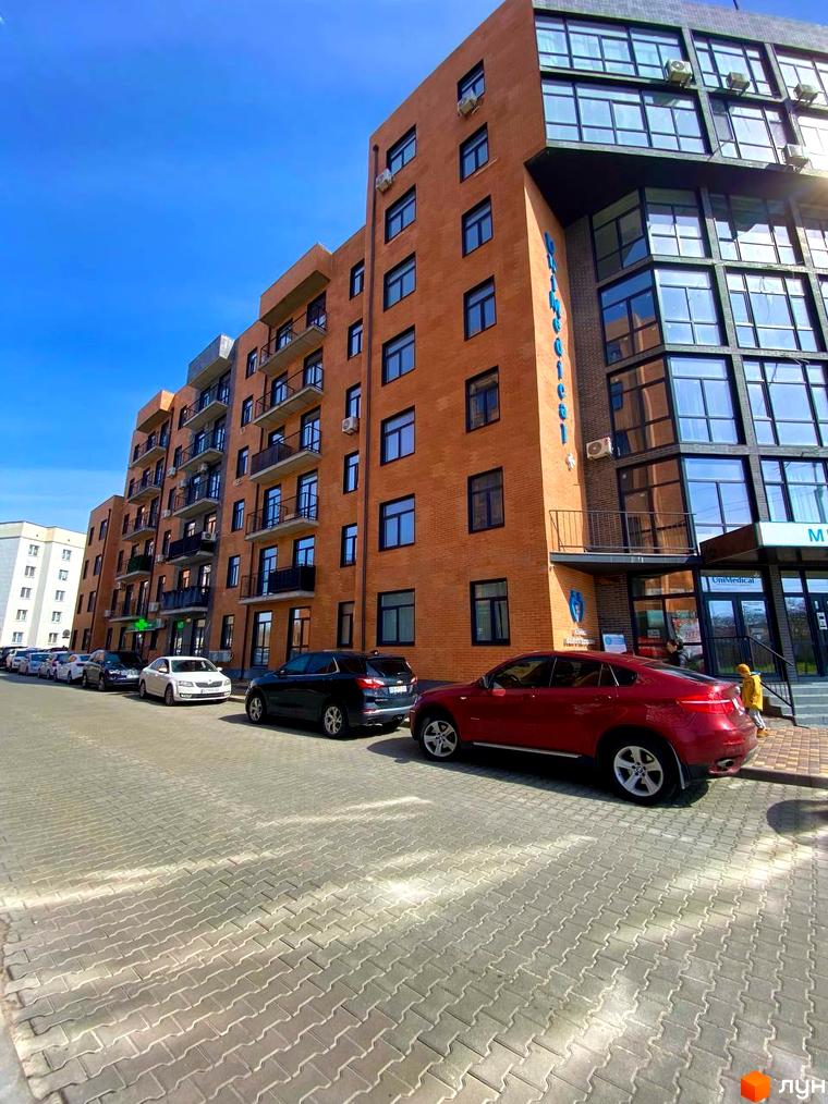 Житловий комплекс Plaza Kvartal з цегляним фасадом та великими вікнами, перед яким розташована парковка з автомобілями.