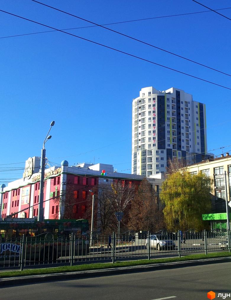 Житловий комплекс Аксіома з сучасним фасадом на фоні чистого неба. Прибудинкова територія з зеленими насадженнями та тротуарами.