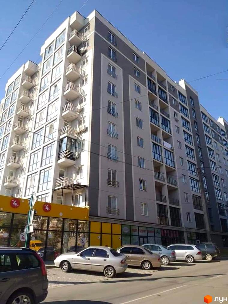 Висотна будівля житлового комплексу з балконами та великими вікнами. Перед будинком припарковані автомобілі, а на першому поверсі розташовані комерційні приміщення.