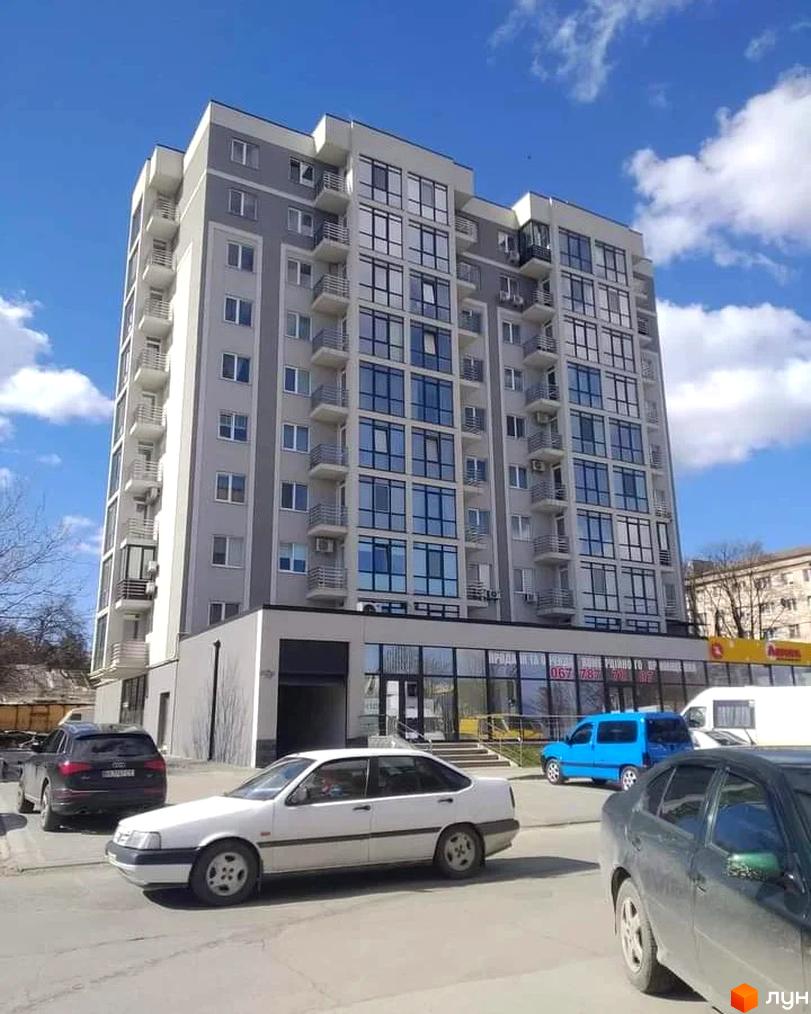 Висотна будівля житлового комплексу з великими вікнами та балконами. На прибудинковій території припарковані автомобілі.