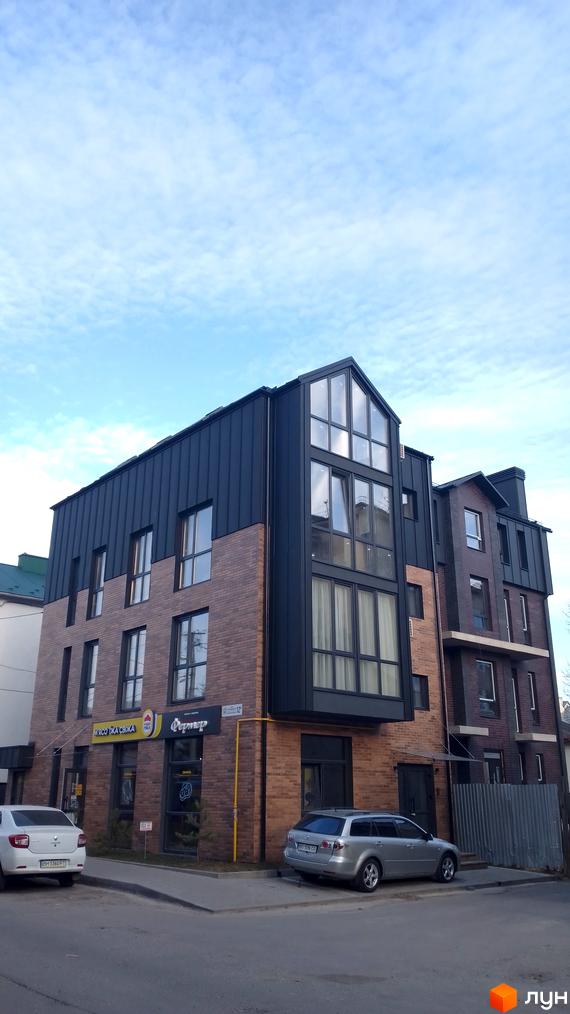 Багатоквартирний будинок з темно-цегляним фасадом та металевою покрівлею, великі вікна. На першому поверсі комерційні приміщення, поруч вулиця з автомобілями.