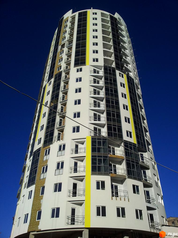 Житловий комплекс Аксіома – висотна новобудова з білою фасадною частиною, жовтими та темними скляними вставками. Будинок має багато балконів та вікон.