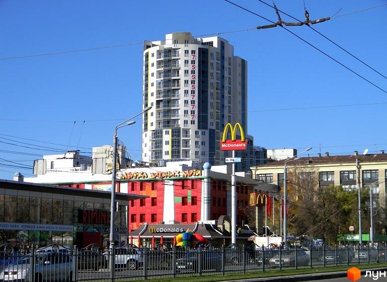 Висотка ЖК Аксіома з фасадом білого кольору та жовтими та сірими елементами. Навколо будівлі видно вивіски McDonald's та інші комерційні об'єкти.