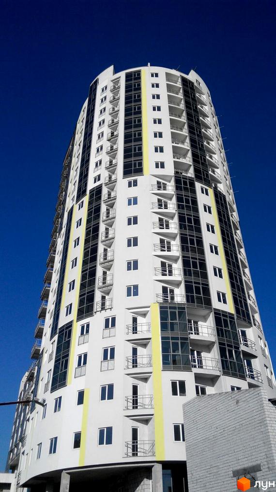 Фасад житлового комплексу Аксіома з білими стінами, темними вікнами та балконами, на тлі синього неба. Будинок має вигнуту форму та жовті вертикальні вставки.