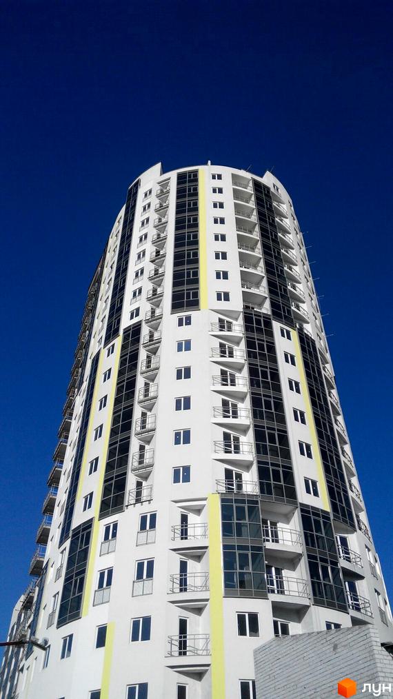 Фасад житлового комплексу Аксіома з численними балконами та панорамним склінням на тлі ясного синього неба. Будинок має біло-сіру кольорову гаму з яскравими жовтими вертикальними елементами.