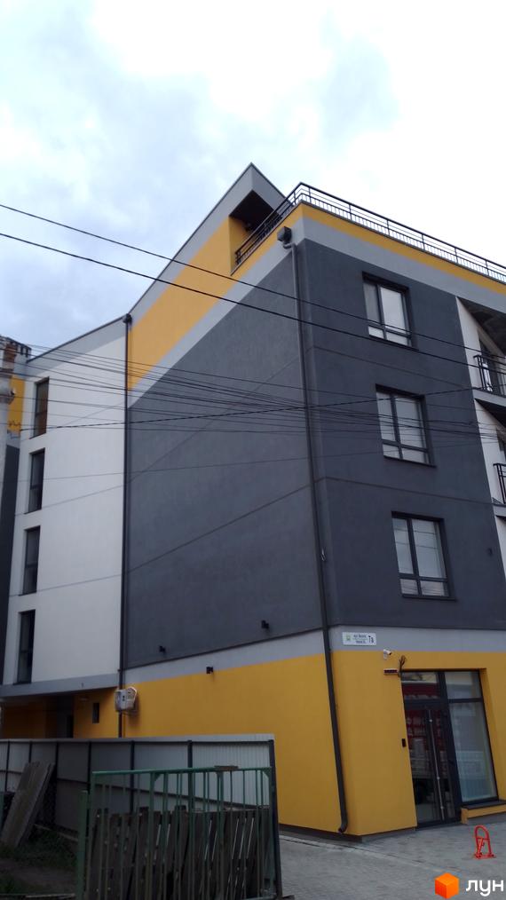 Багатоповерховий будинок з жовтими та сірими фасадами. На першому поверсі розташовані комерційні приміщення. На території є паркан.