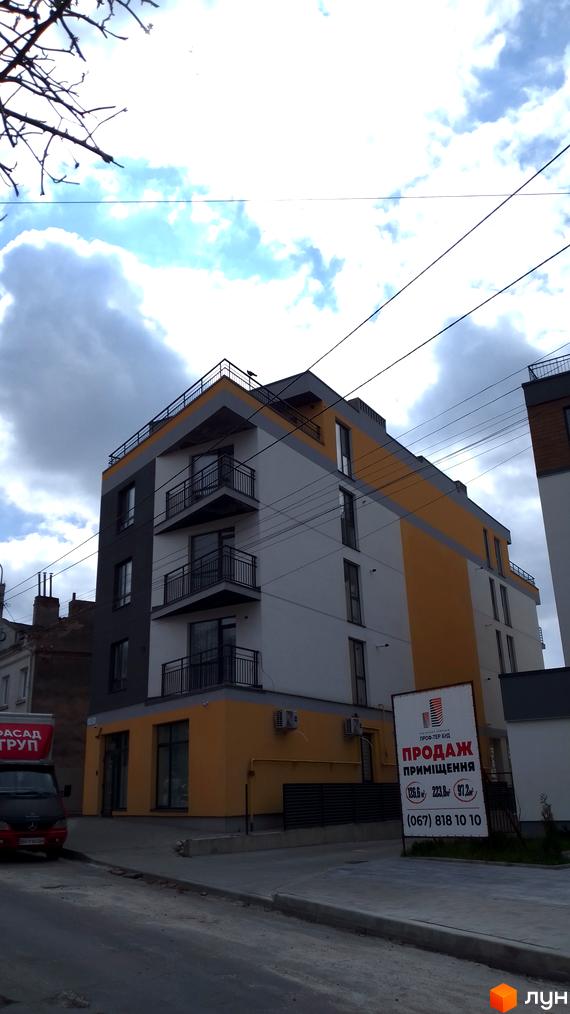 Багатоповерховий будинок з балконами та жовтим фасадом. На передньому плані асфальтована дорога та рекламний щит з інформацією про продаж приміщень.