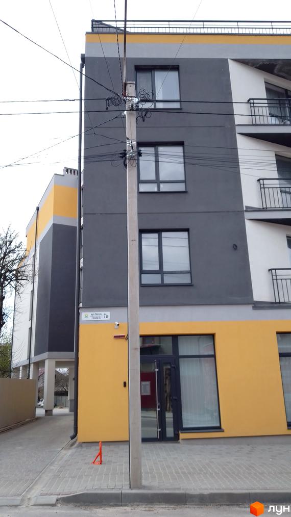 Багатоповерховий будинок з сіро-жовтим фасадом та балконами на вул. Весела, 7. Перед ним тротуар та стовп з дротами.