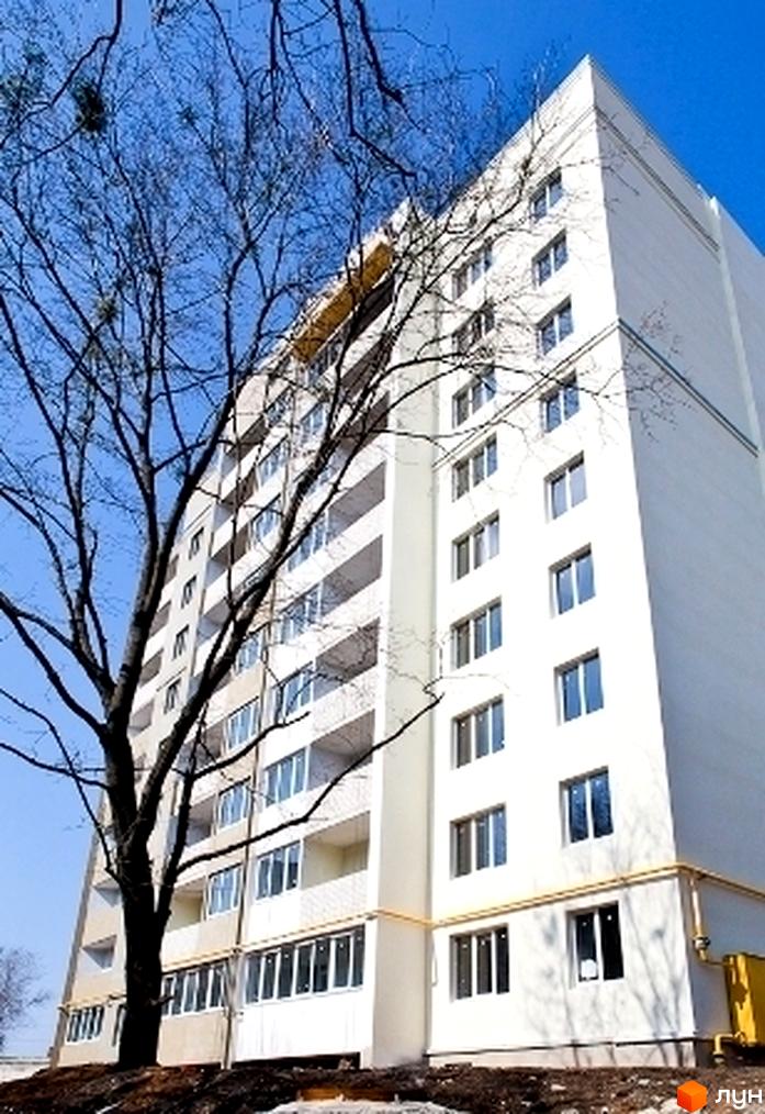 Зовнішній вигляд новобудови на просп. Олександрівський (Косіора), 69е. Багатоповерховий будинок з білим фасадом та балконами. Навколо будівлі дерева та ділянка з ґрунтом.