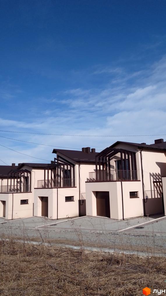 Бежеві будинки з коричневими дахами та балконами. Територія перед будинками викладена плиткою, видно суху траву.