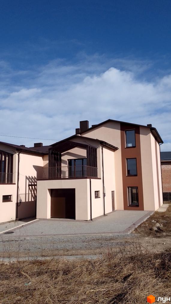 Двоповерховий будинок з балконом та гаражем. Фасад будинку пофарбований у світлі тони, поруч вимощена бруківкою прибудинкова територія.