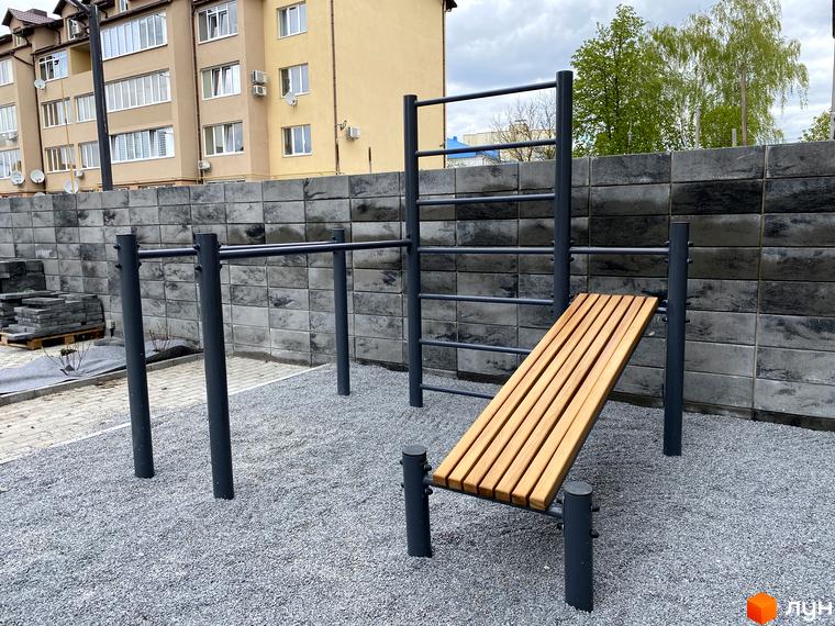 Клубний будинок „Васильченка“ з сірою цегляною стіною та зоною для тренувань на подвір'ї. Навколо будинку розташована бруківка та гравійне покриття.