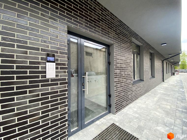 Клубний будинок Васильченка: вхідна група з домофоном та скляними дверима, фасади оздоблені темною цеглою, перед будинком викладена плитка.