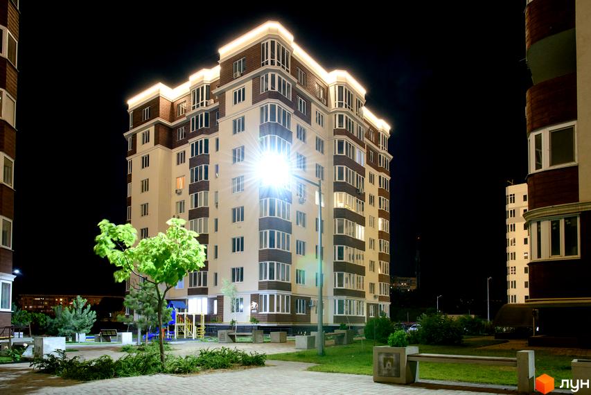 Висотна будівля житлового комплексу Золота Ера вночі з підсвічуванням. На передньому плані зелені насадження та лавки.